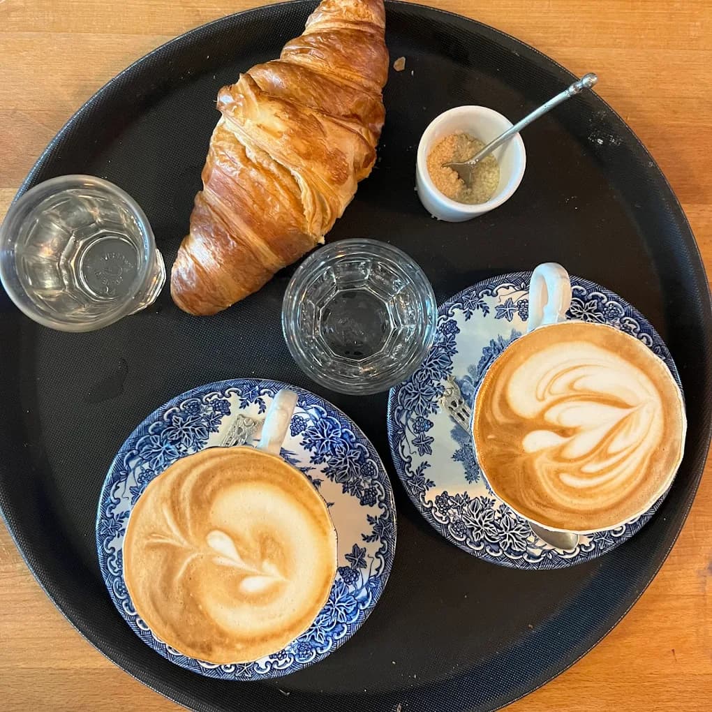 Deux cappuccinos au latte art dans des tasses vintage dépareillées et un croissant, sur un plateau au Café Le Passage.
